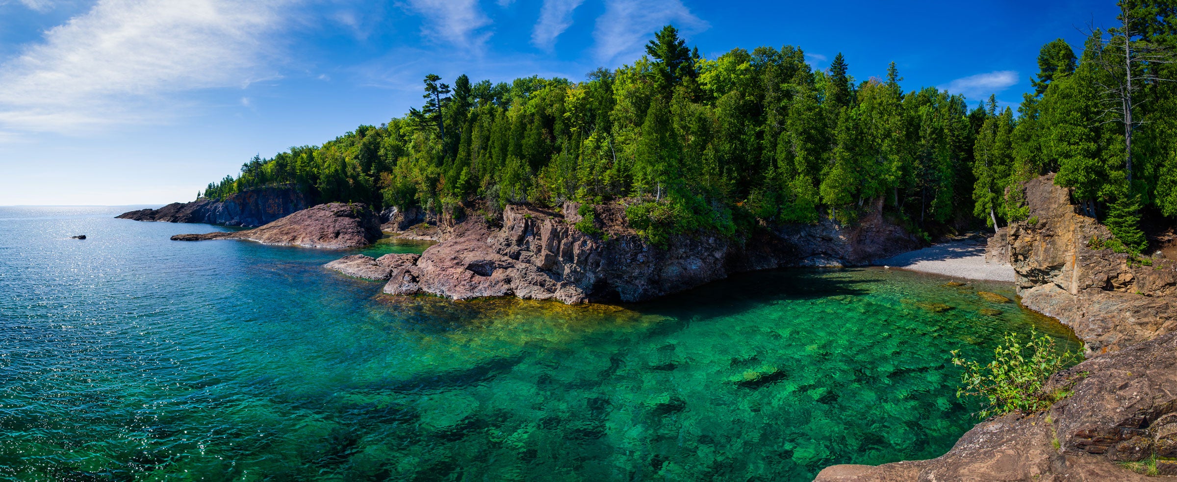 A panoramic photo of a clear body of water next to a mountainous forest.