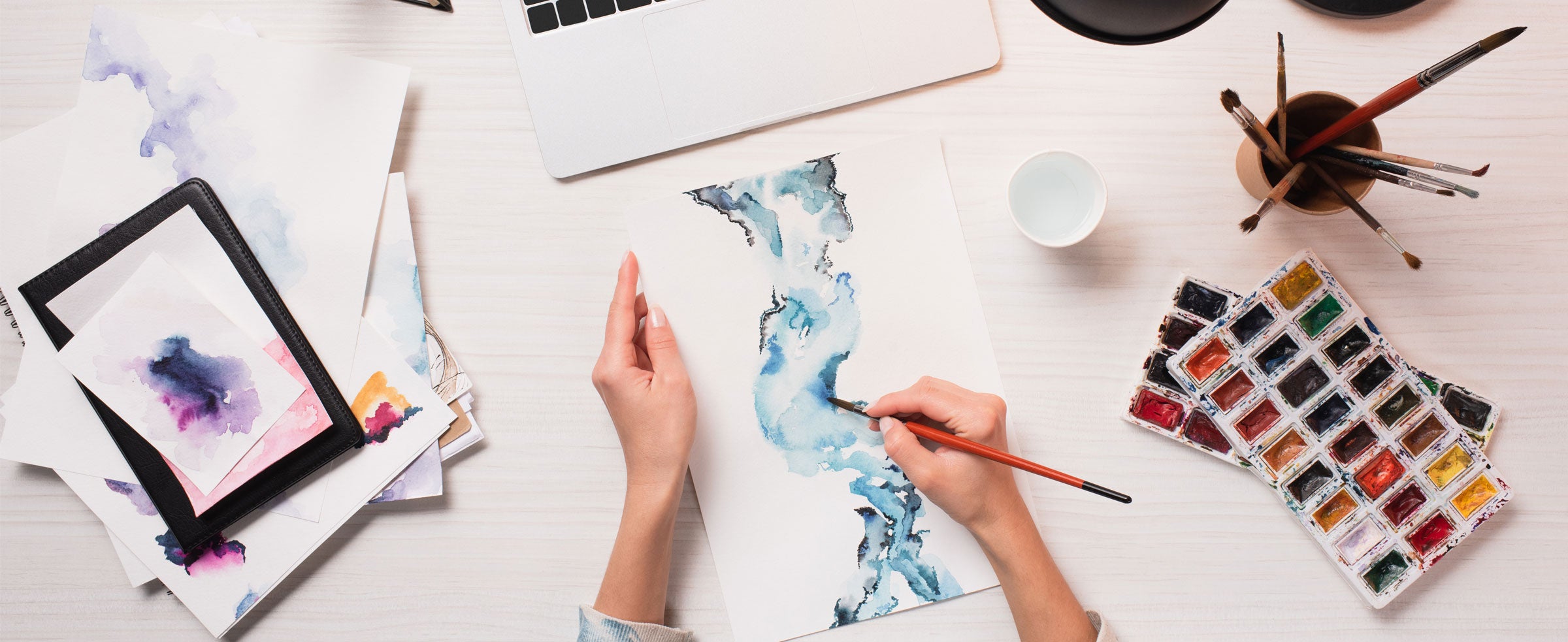 An overhead image of an artist's desk while two hands paint with water colors.. Many creative supplies are visible.