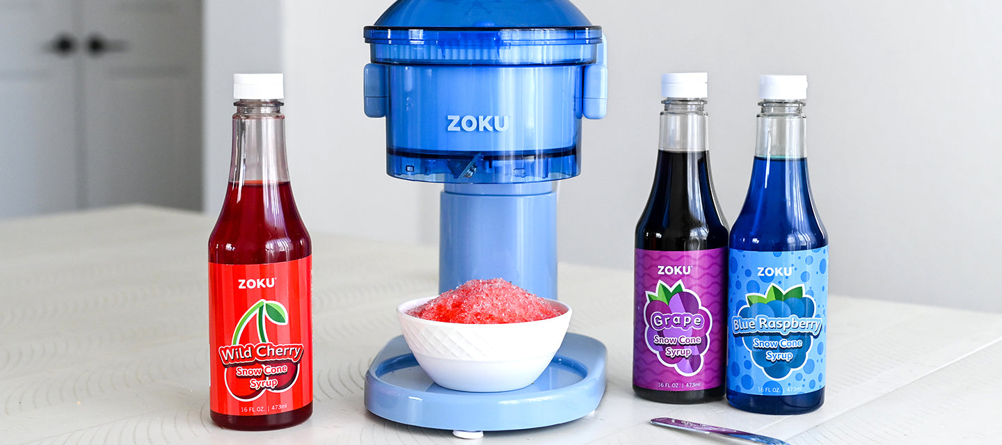 ZOKU Snow Cone Maker with a bowl of shaved ice in front, flanked by bottles of ZOKU Wild Cherry, Grape, and Blue Raspberry Snow Cone Syrups on a white table.
