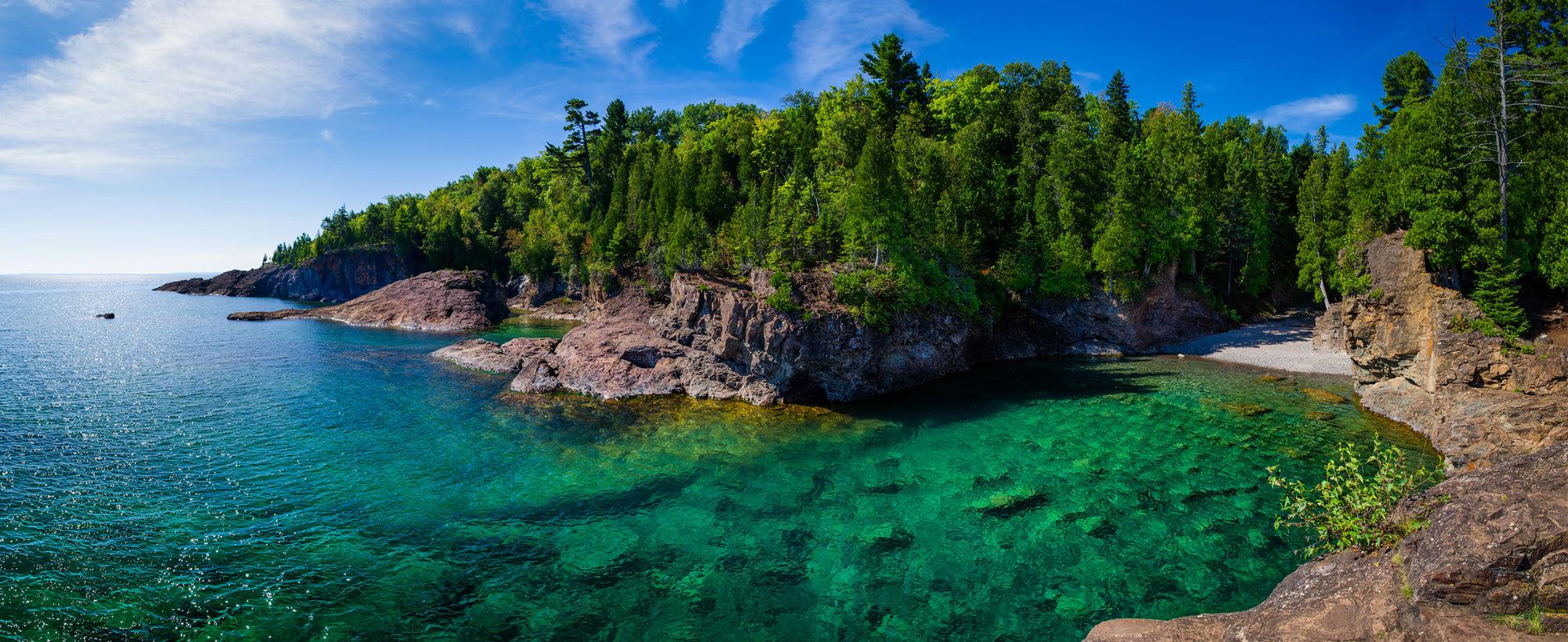 A panoramic photo of a clear body of water next to a mountainous forest.