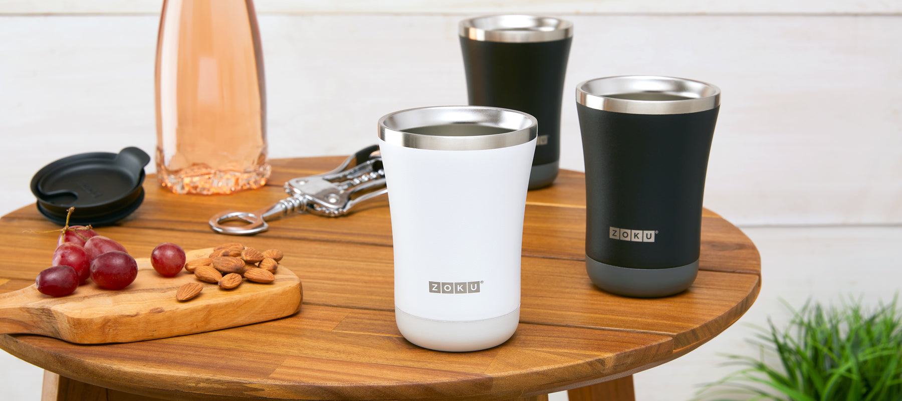  The ZOKU 12oz 3-in-1 Tumbler in Black and White elegantly displayed on a wooden table. Surrounding it is a spread of grapes, nuts, a bottle opener, and a wine glass in the backdrop.