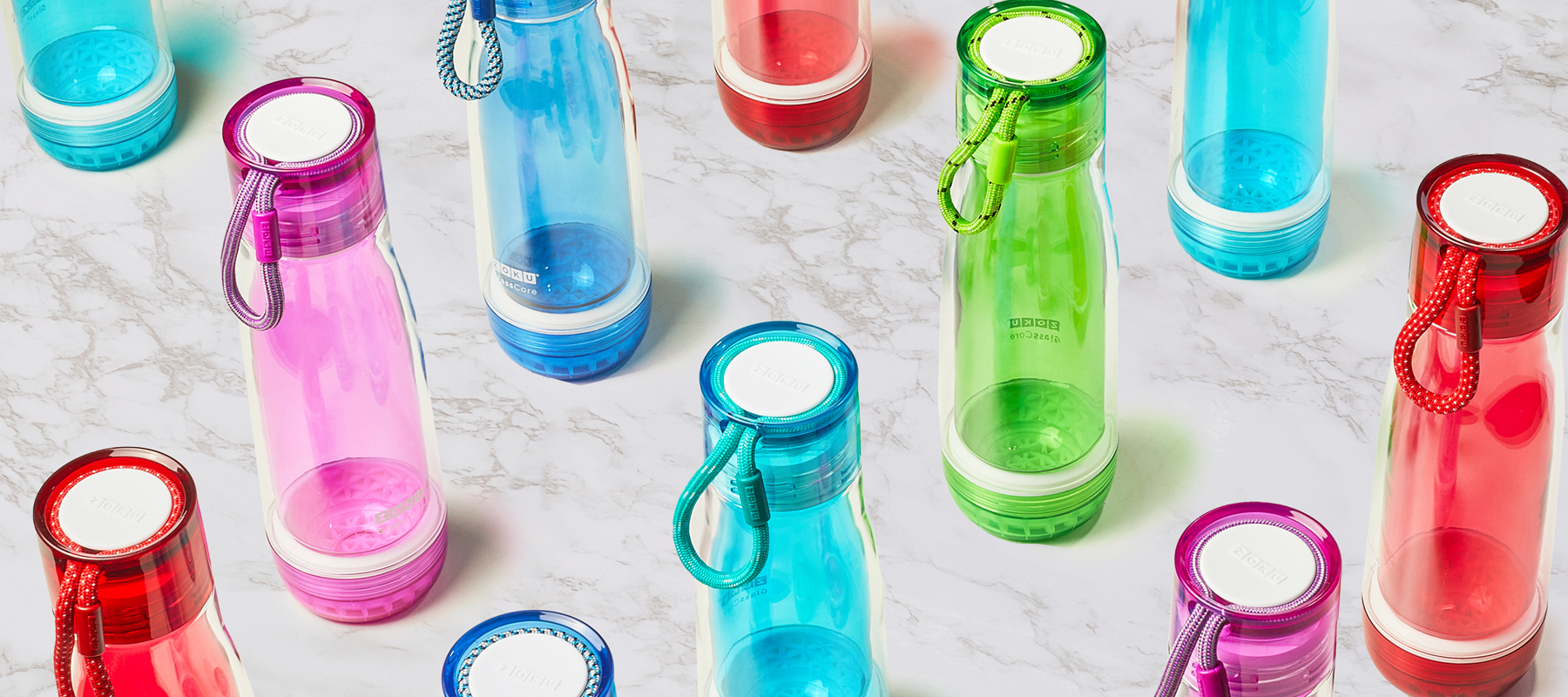  An assortment of the ZOKU 16 oz Glass Core Bottles in the colors Light Blue, Blue, Green, Red, and Purple displayed on a sleek, white marble table.